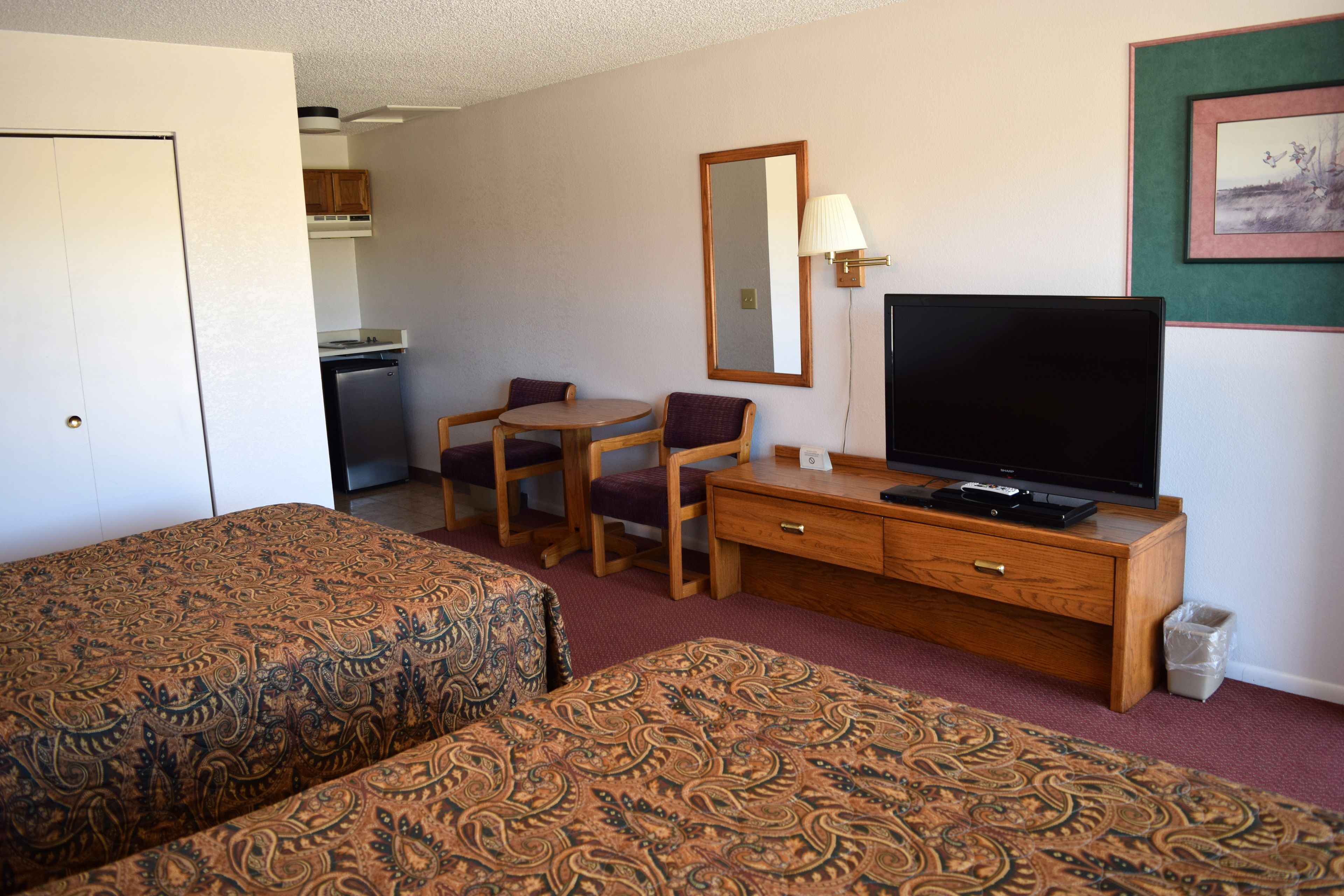 A hotel room with two beds, a desk, a television, and a small kitchenette.