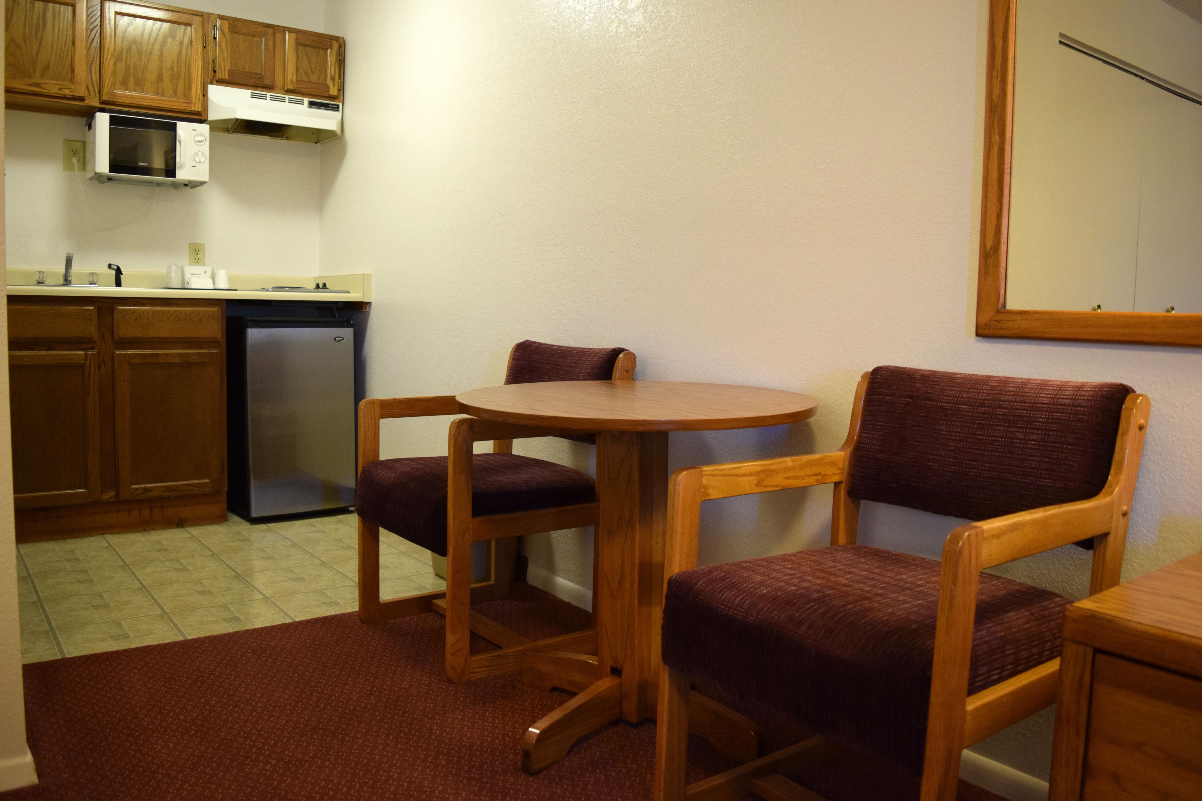 A small kitchen with wooden cabinets, a microwave, and a refrigerator. There is a round table with two chairs and a rug on the floor.