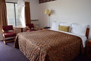 A hotel room with a large bed, a small table, two chairs, a lamp, and a view of a staircase.