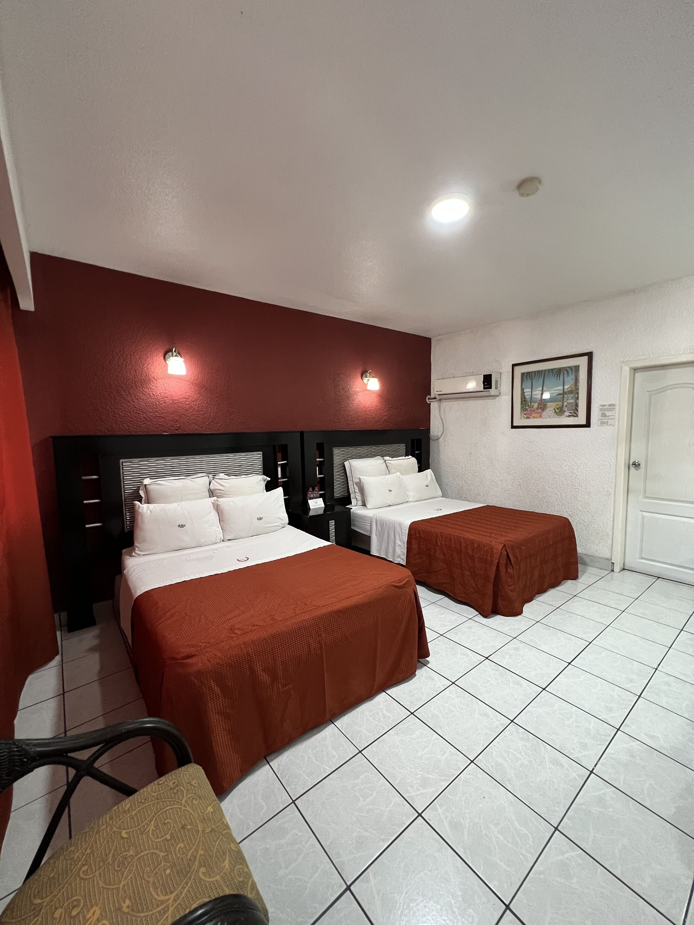 Habitación de hotel con dos camas, una pared de acento roja, una puerta blanca y un cuadro en la pared.
