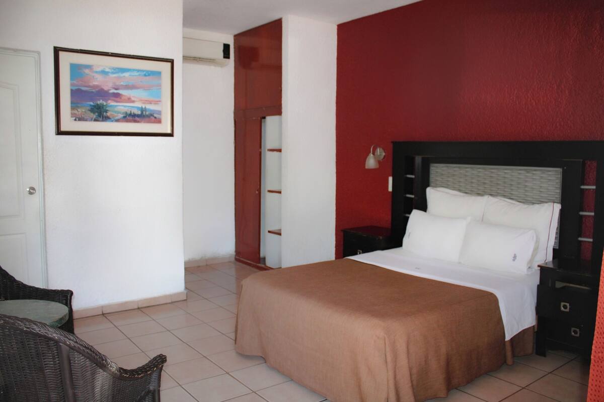 A hotel room with a bed, a chair, a nightstand, a lamp, and a painting on the wall.