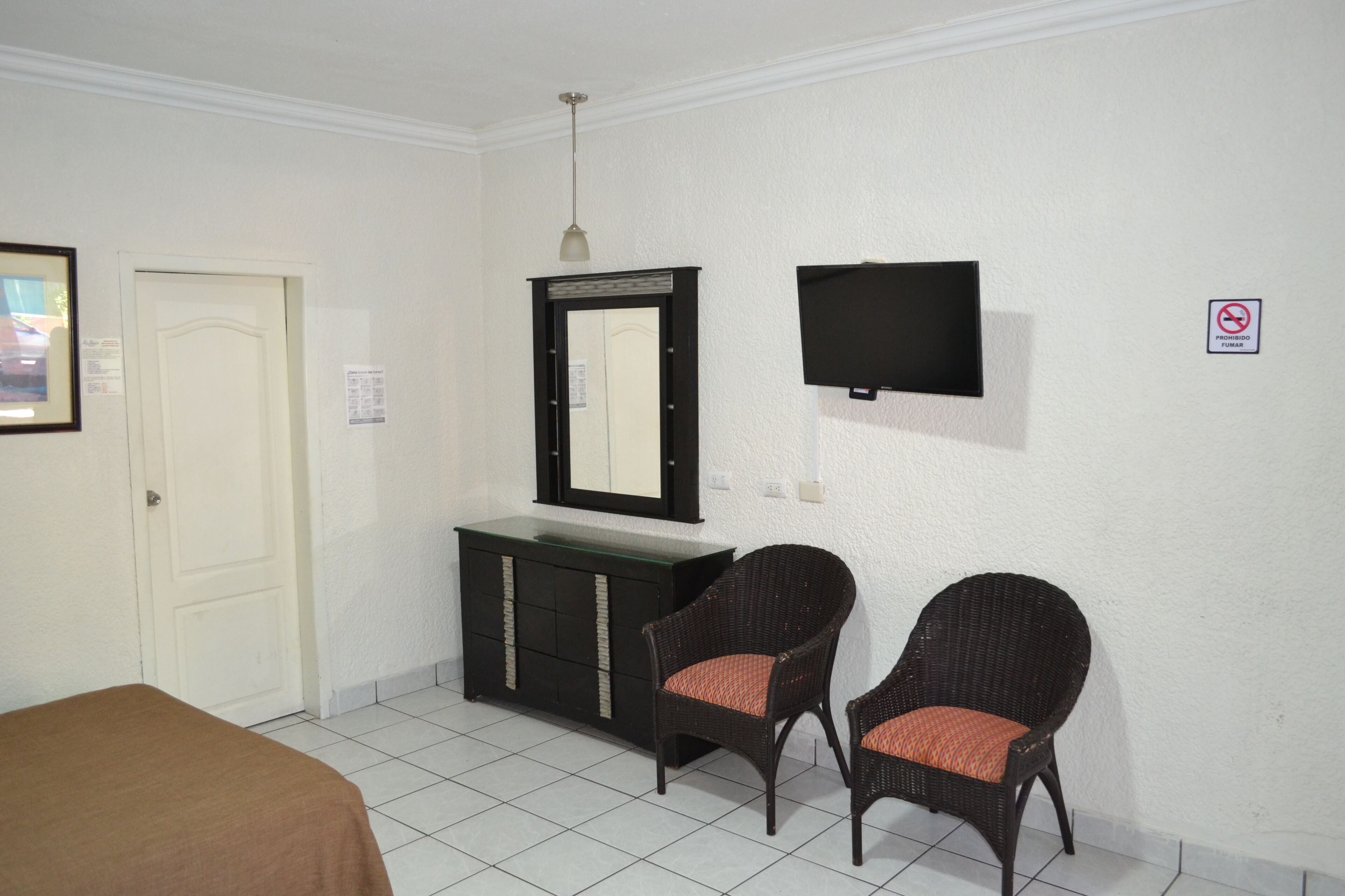 Une chambre d’hôtel avec un lit, une commode, deux fauteuils en osier, un téléviseur et une porte.
