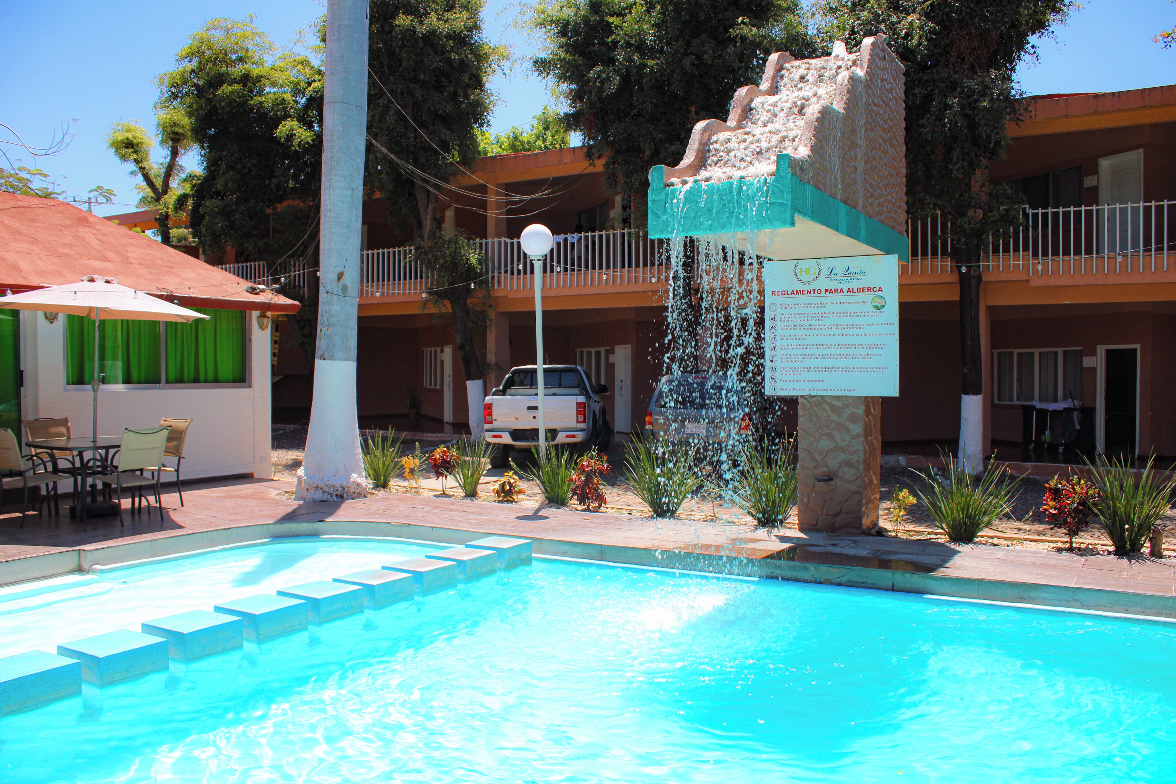 Una piscina con fuente de agua, mobiliario exterior y un edificio con balcones al fondo.