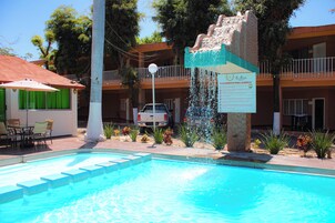 Una piscina con fuente de agua, mobiliario exterior y un edificio con balcones al fondo.