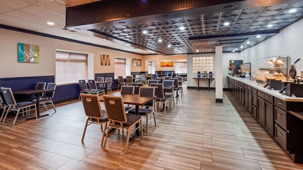 A spacious dining area with multiple tables and chairs, a kitchen area with appliances, and a ceiling with a grid pattern.
