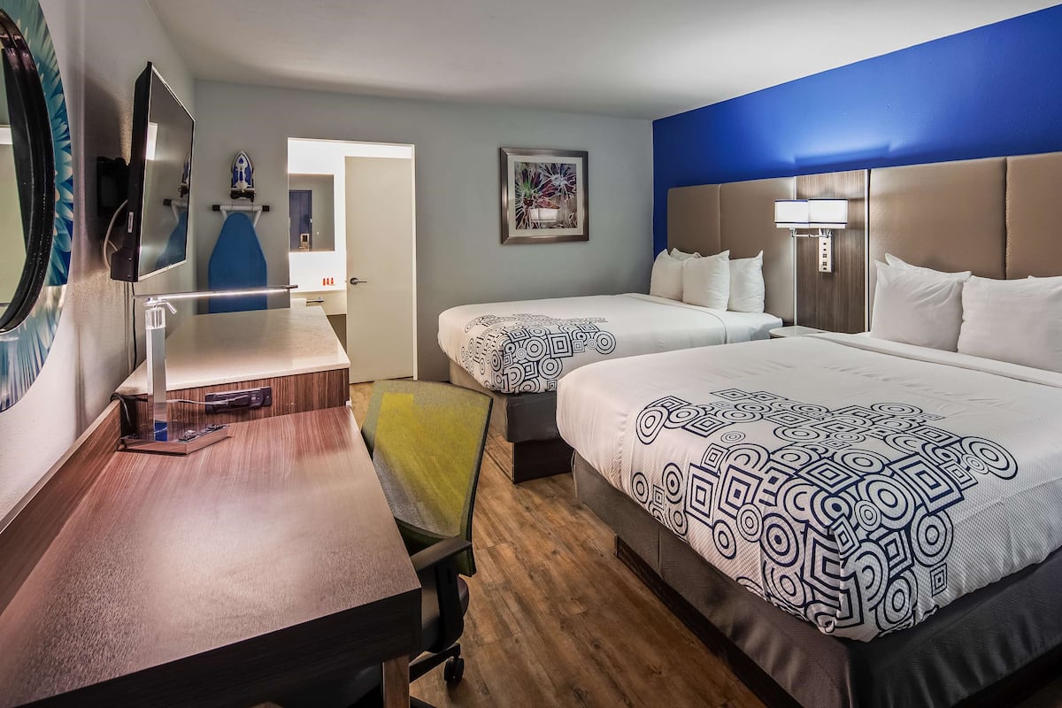 A hotel room with two beds, a desk, a chair, a TV, and a bathroom visible through an open door.