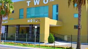 A hotel named "TWO" with a modern yellow facade, large windows, and palm trees in the foreground.