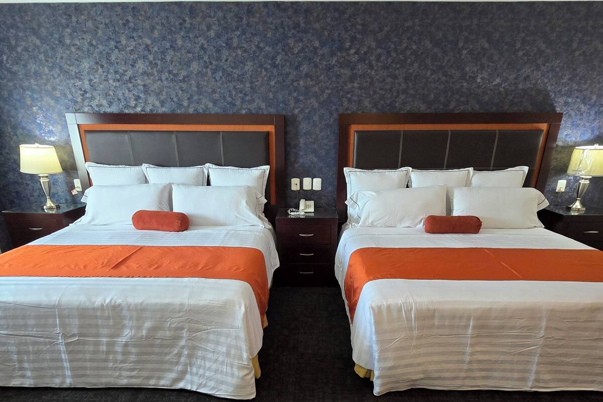A hotel room with two beds, each with white bedding and orange accents, against a dark blue textured wall.