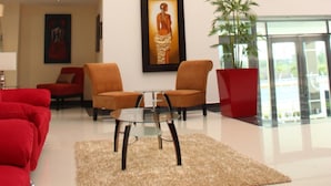 A spacious lobby with a high ceiling, featuring a modern seating area with red sofas and armchairs, a glass coffee table, and a decorative rug.