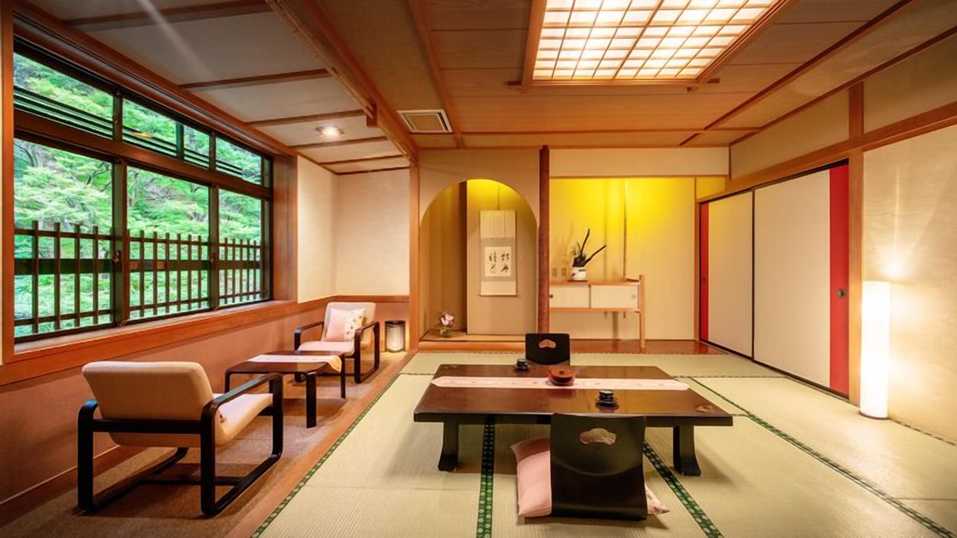 A traditional Japanese room with tatami flooring, a low table, chairs, a sliding door, and a view of greenery through large windows.