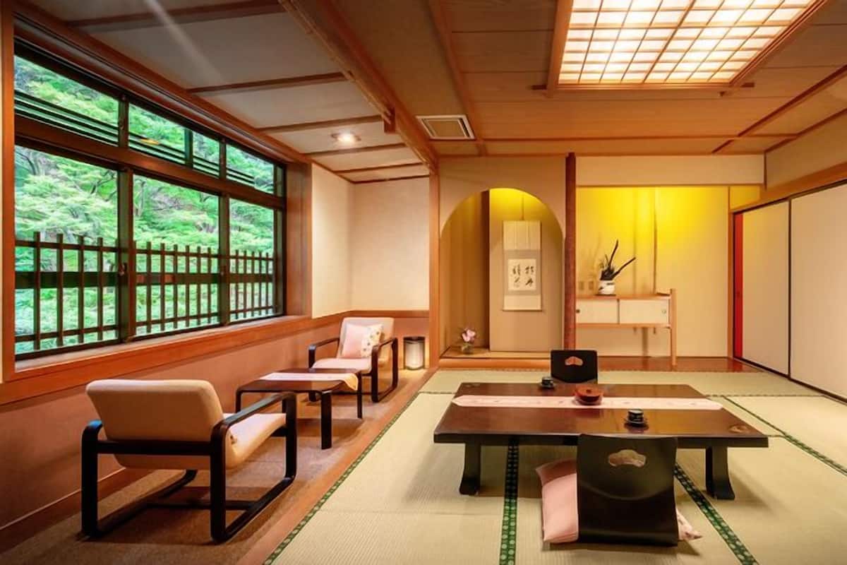 Une pièce de style japonais traditionnel, avec un sol recouvert de tatamis, une table basse, des chaises, une porte coulissante et une vue sur la verdure grâce à de grandes fenêtres.