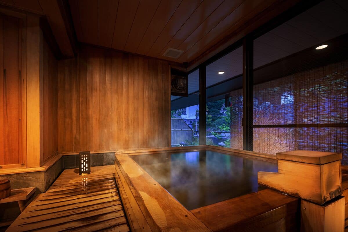 Un onsen japonais traditionnel, avec un intérieur en bois, un bain d’eau chaude et une vue sur l’extérieur grâce à de grandes fenêtres.