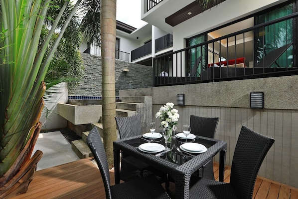 An outdoor dining area with a table set for four, surrounded by wicker chairs, a stone wall, and a palm tree.