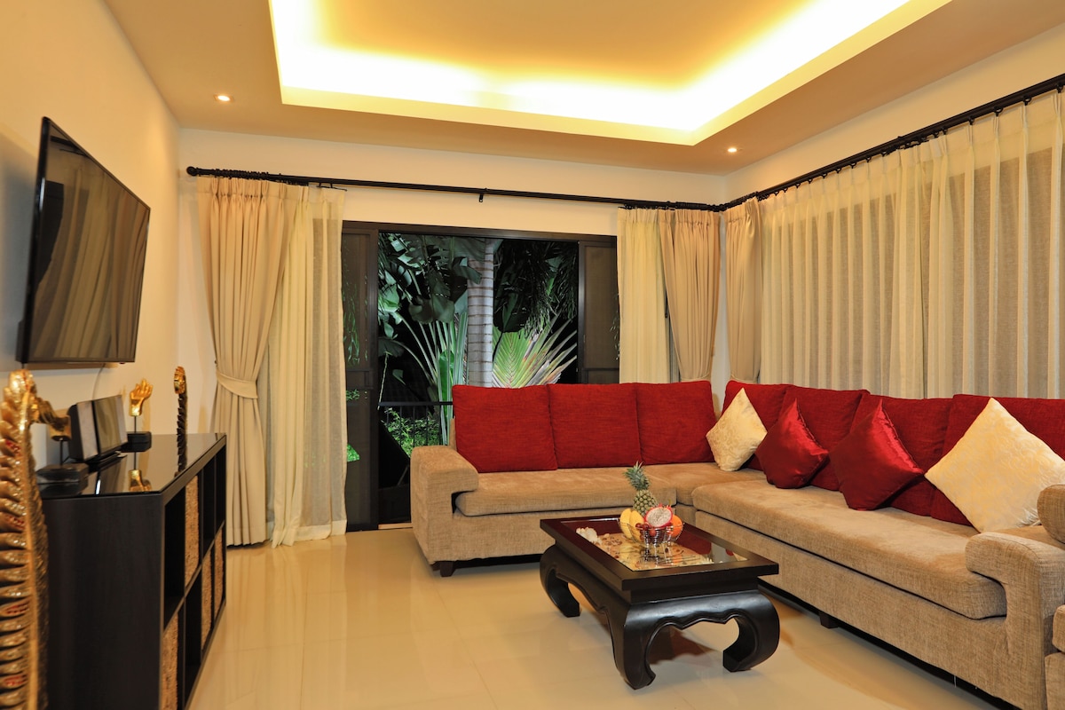 A living room with a beige sofa, red cushions, a coffee table with fruit, and a TV on the wall.