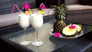 Dos vasos de una bebida blanca con pajitas rosas y adornos, una piña y una mesa de mimbre.