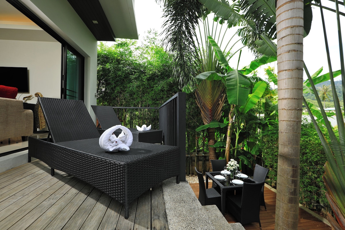 A modern outdoor lounge area with a black wicker sofa, a dining table with chairs, and lush tropical plants.