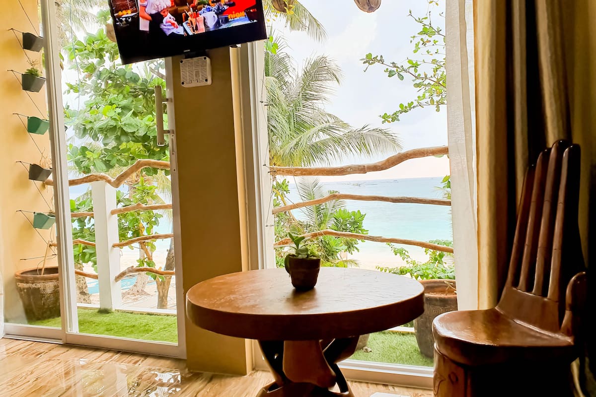 A room with a TV on the wall, a round wooden table, and a view of the ocean through large windows.