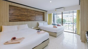 A hotel room with two beds, a wooden headboard, a large window with curtains, and a balcony with a view of buildings. - Royal Beach Residence (Patong)