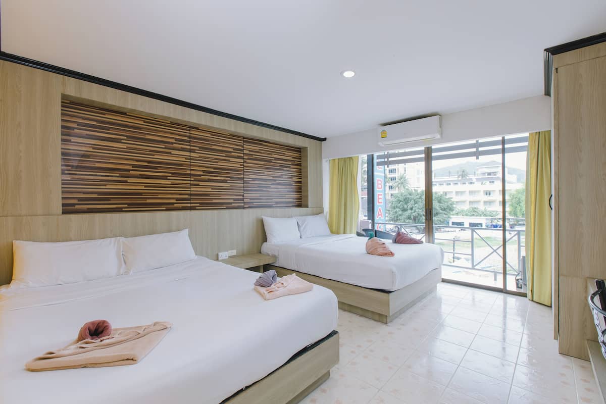 A hotel room with two beds, a wooden headboard, a large window with curtains, and a balcony with a view of buildings.