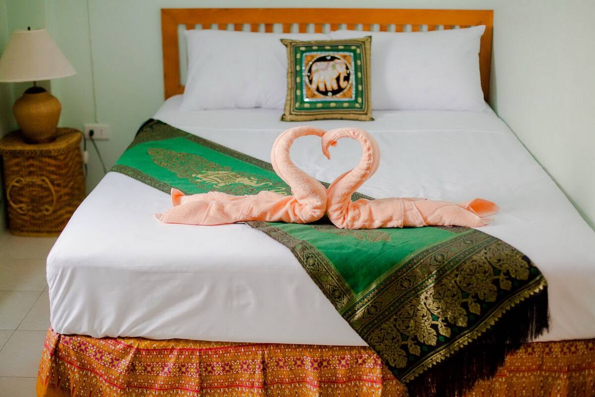 Una cama bien arreglada con una toalla dispuesta en forma de corazón, una colcha verde y dorada, y una almohada decorativa.