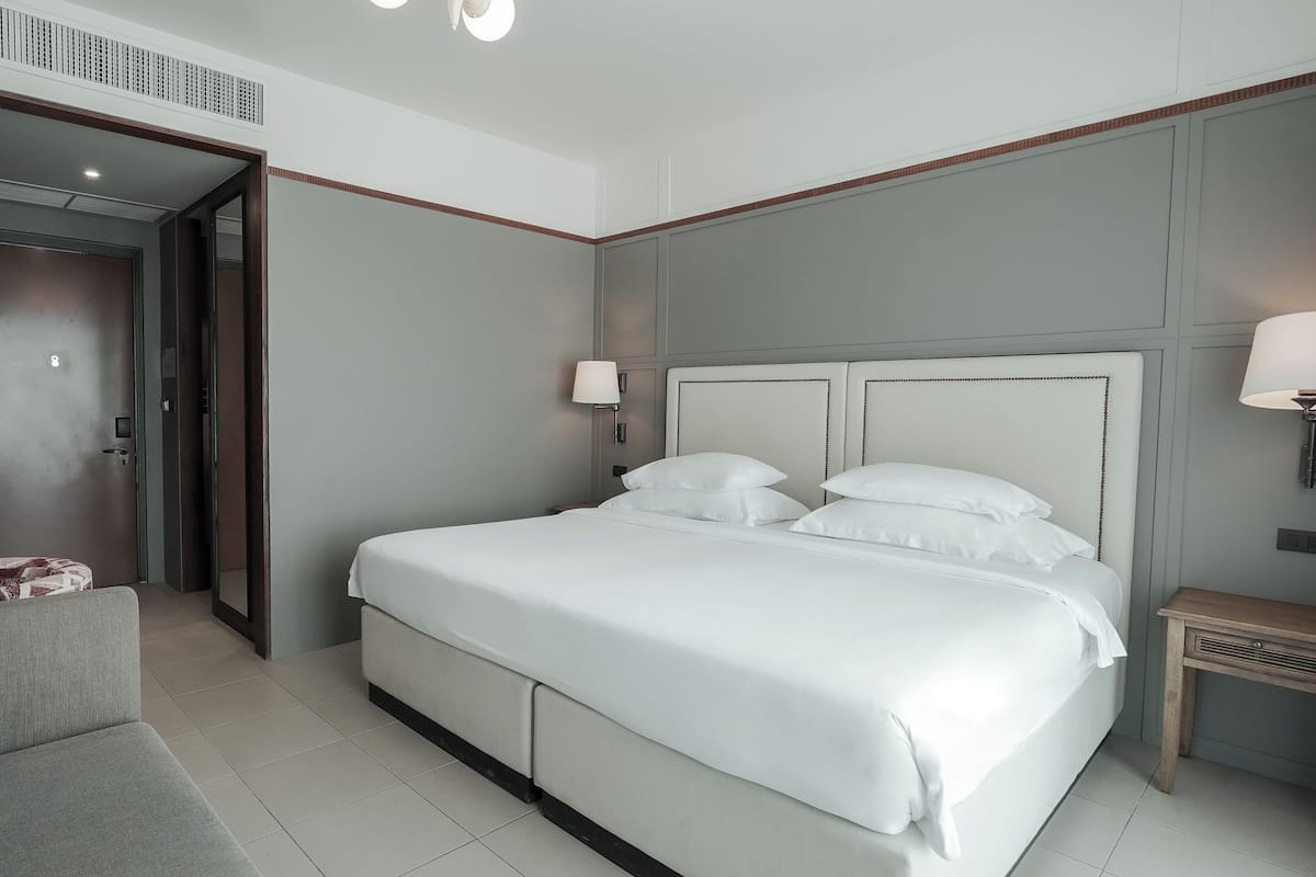 Habitación de hotel con una cama grande, un televisor montado en la pared, una mesita de noche con lámpara y un sofá con cojín escocés.