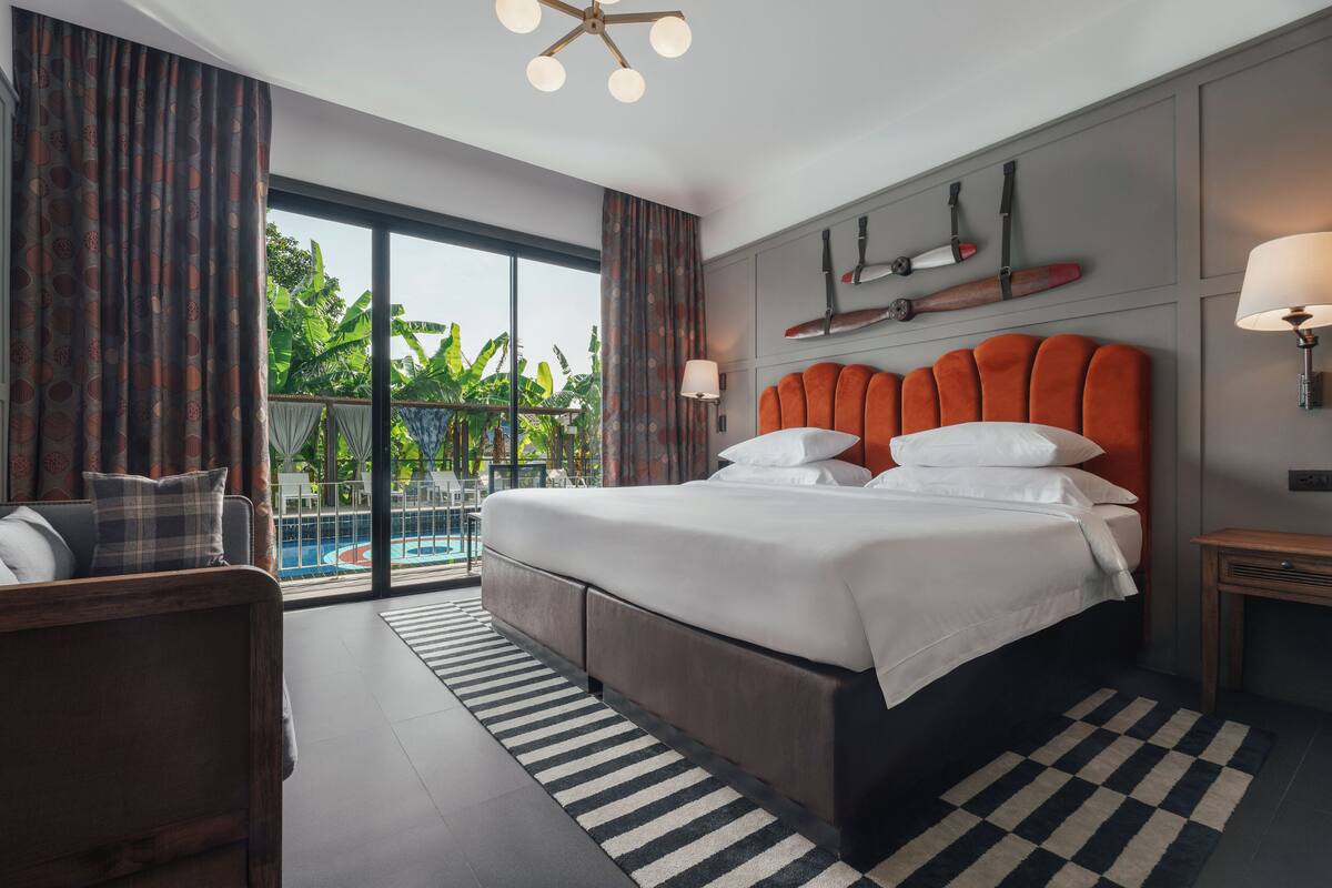 Una habitación de hotel moderna con una cama grande, un sofá, vista a una piscina y un balcón con plantas.