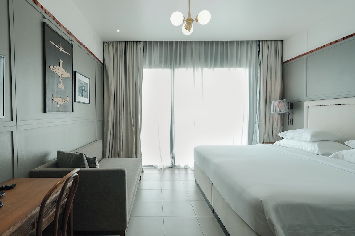 Habitación de hotel con una cama grande, un escritorio con teléfono, una silla y un ventanal con cortinas.