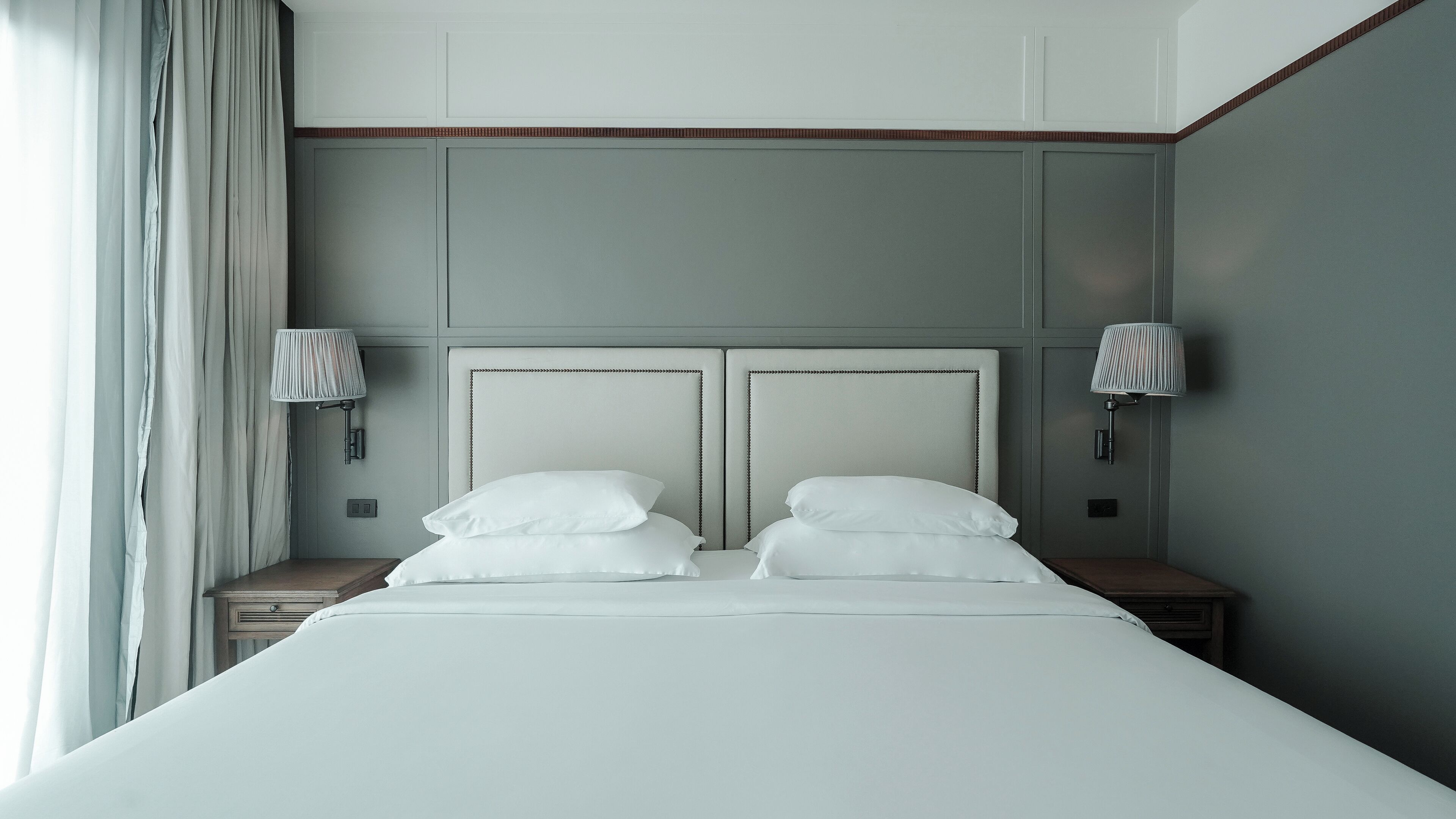 A neatly made bed with white linens and pillows, two bedside tables with lamps, and a large window with curtains.
