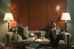 Two individuals seated on a sofa in a room with a wooden panel wall, two lamps, and a small table with books and a camera.