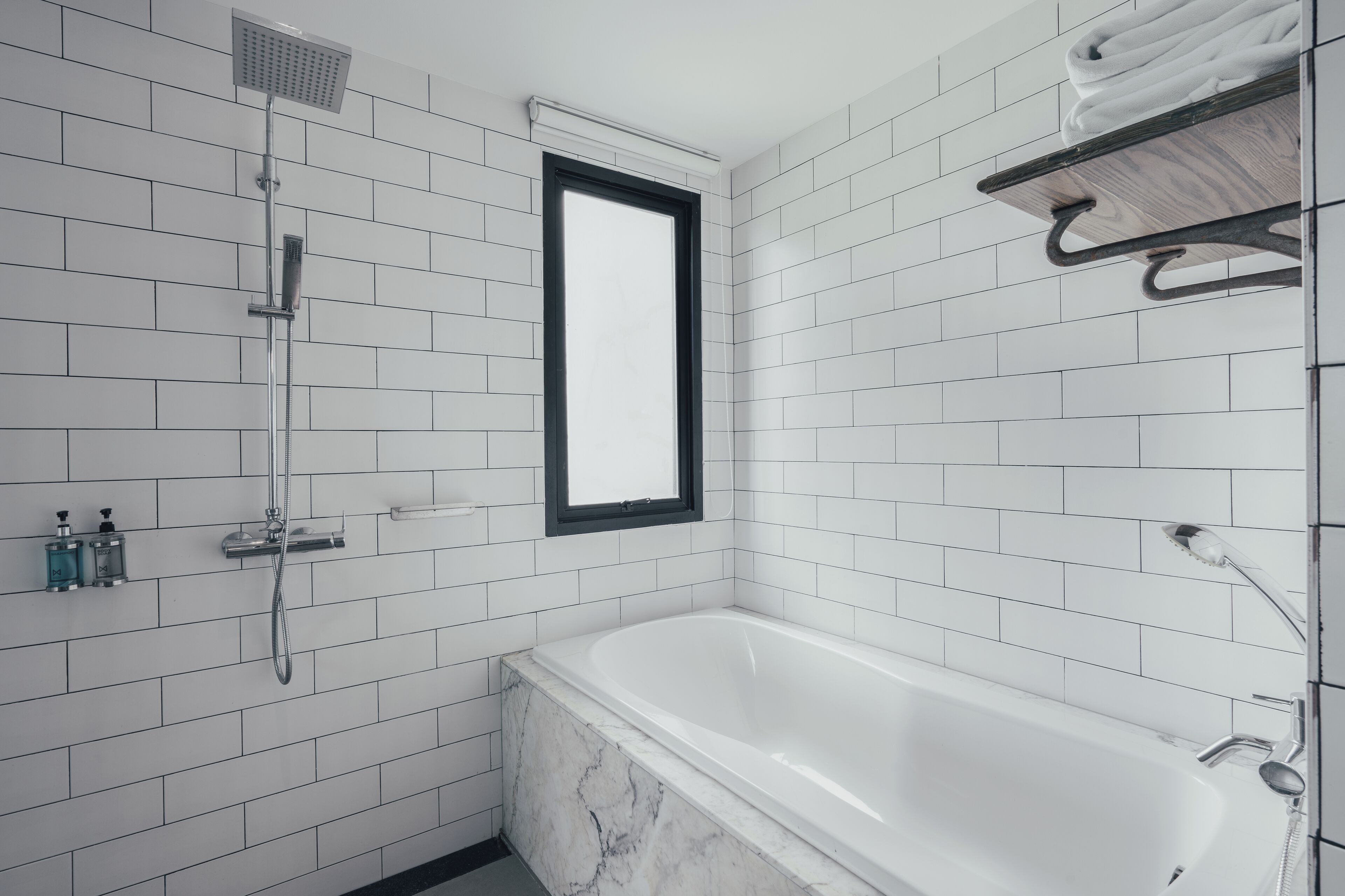 A modern bathroom with white tiled walls, a bathtub, a showerhead, and a window.
