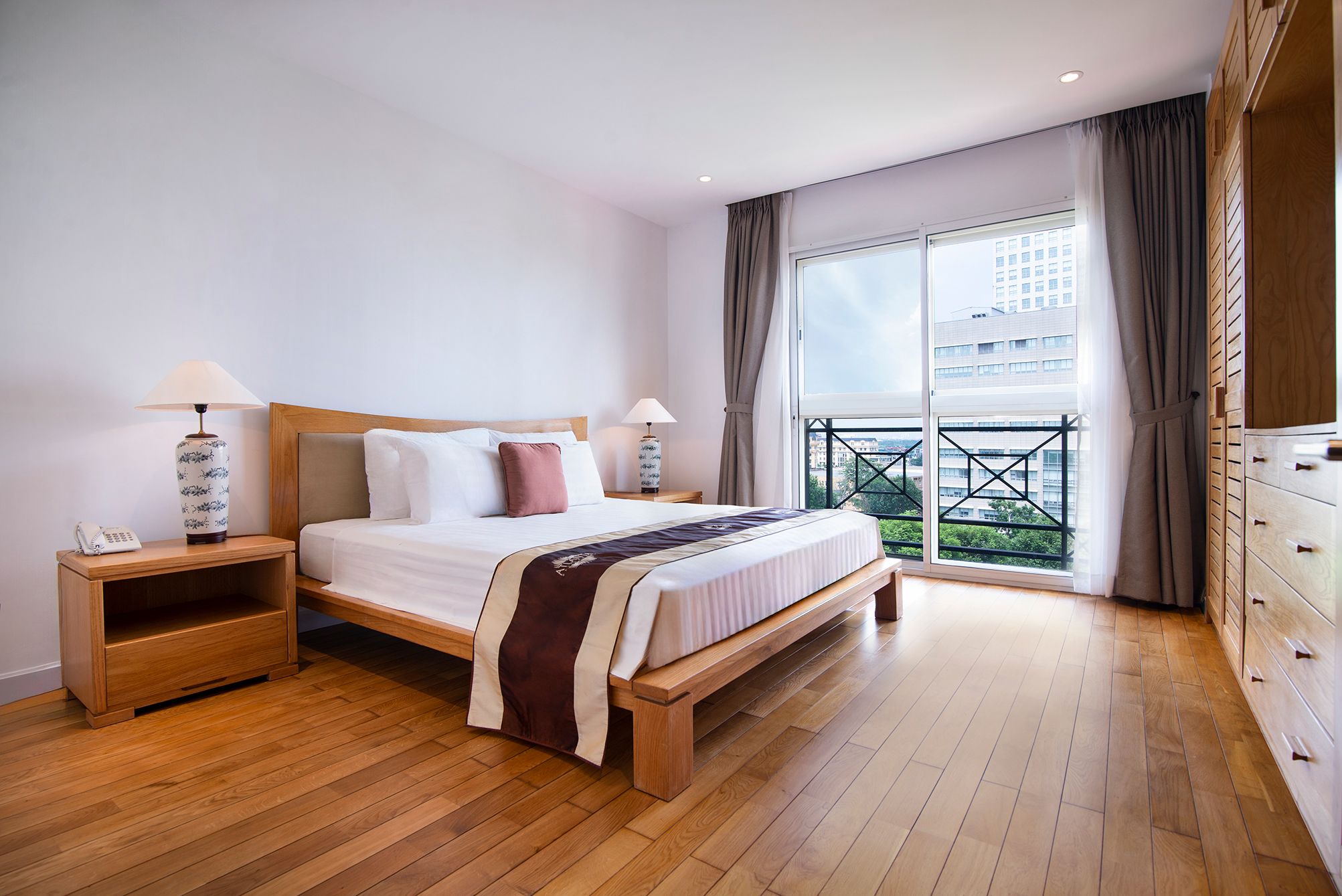 A hotel room with a wooden bed, bedside tables, a wardrobe, and a balcony with a city view.