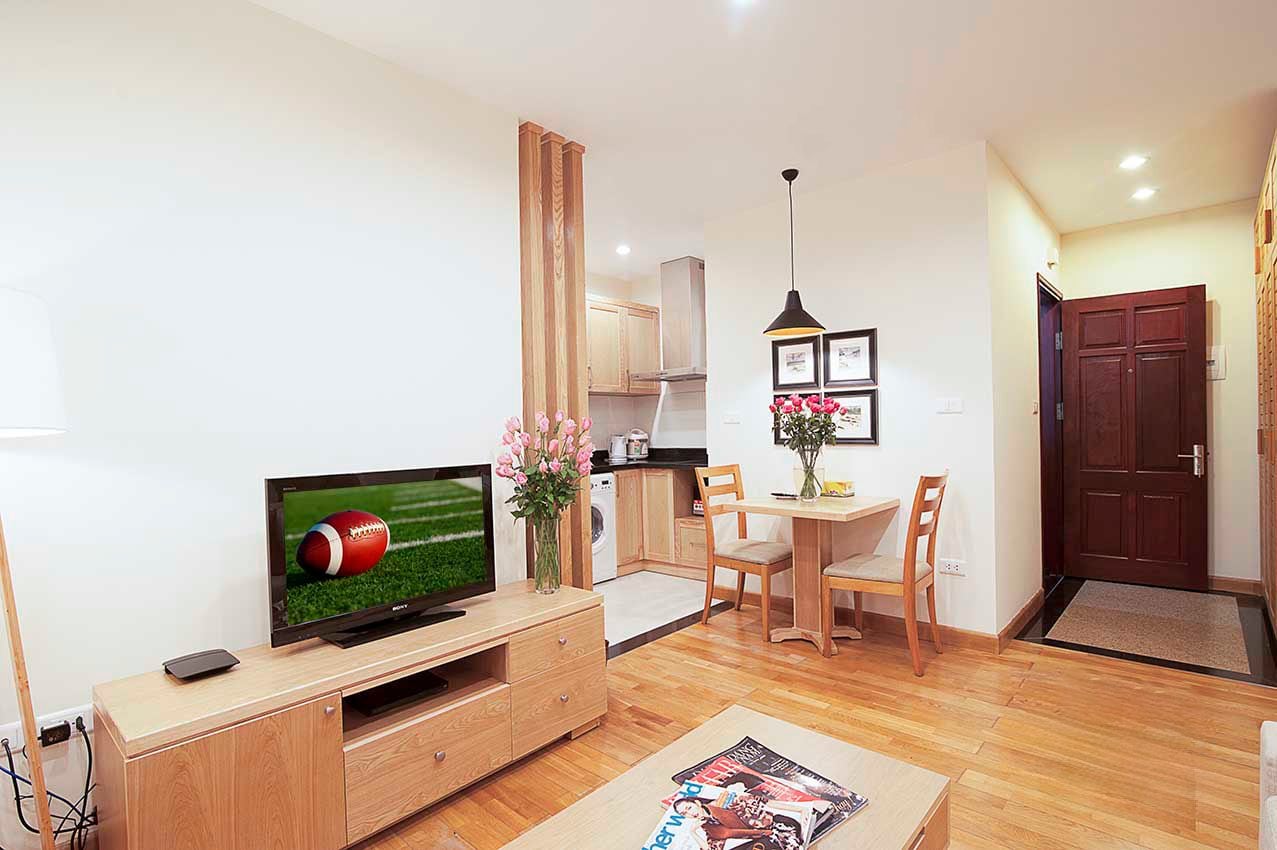 A modern living room with a flat-screen TV, wooden furniture, a dining area, and a kitchen in the background.