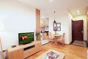 A modern living room with a flat-screen TV, wooden furniture, a dining area, and a kitchen in the background.