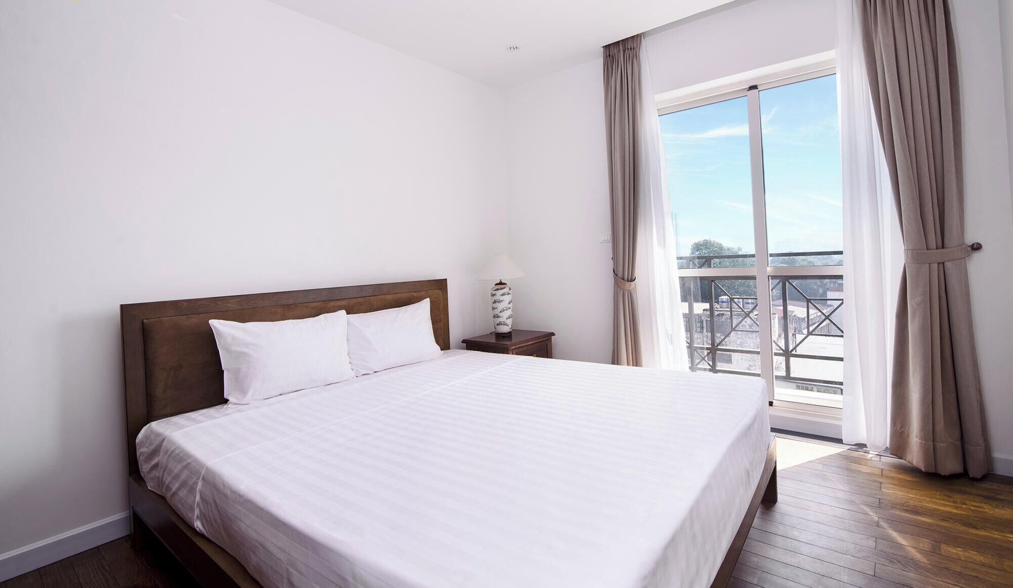 A bedroom with a large bed, a nightstand, a lamp, and a view of a balcony through a window.