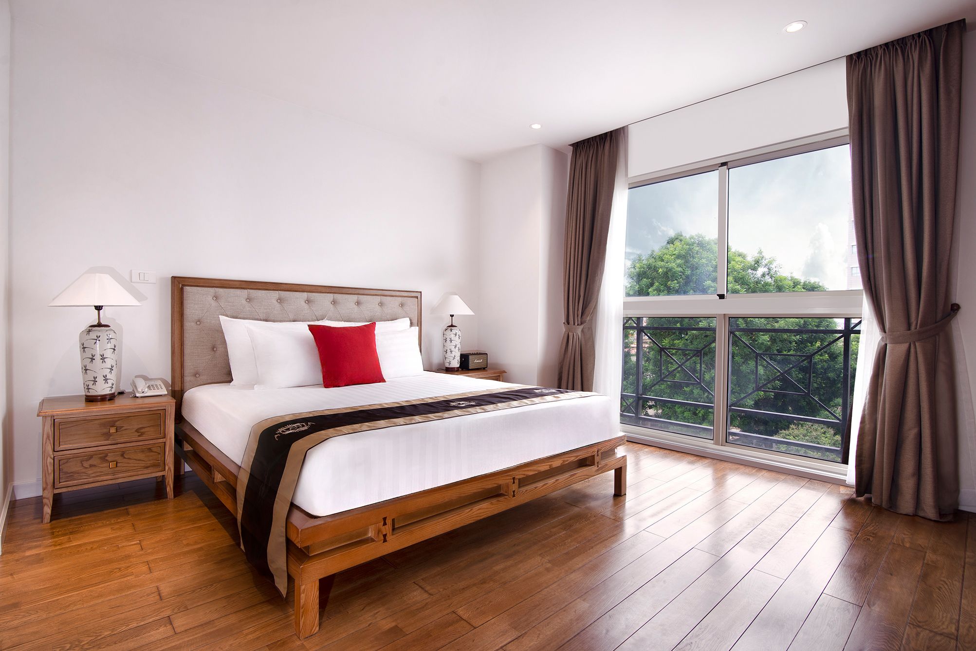A bedroom with a wooden bed, white bedding, a red pillow, two bedside lamps, a nightstand, and a large window with curtains.