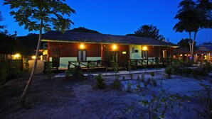 A wooden building with a thatched roof, surrounded by small trees and a sandy ground. - PP Red Tuna Hut (Ko Phi Phi)