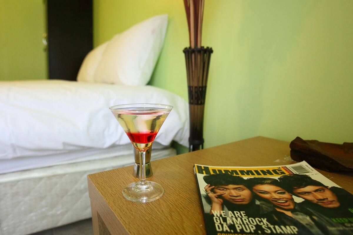 A martini glass on a wooden table with a magazine featuring a group photo. A bed with white bedding and a green wall is in the background.