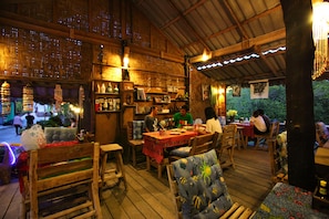 Un interior rústico de madera con mesas y sillas, una zona de bar con botellas y vasos, y vistas a zonas verdes al exterior.
