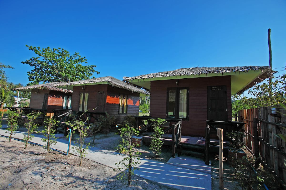 Two rustic wooden houses with thatched roofs, surrounded by young trees and a clear blue sky.