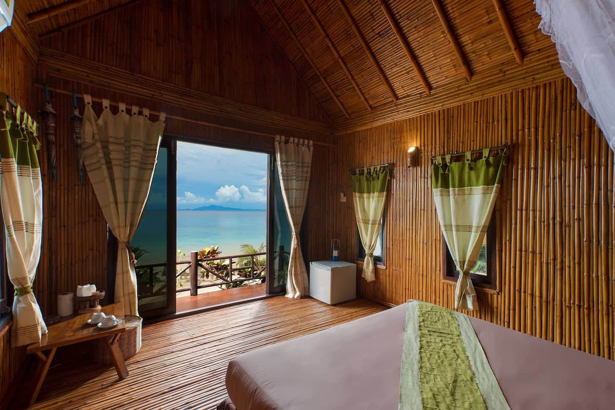 Ein Zimmer mit Holzboden, einem Bett, einem Tisch mit einem Korb und Blick auf das Meer durch große Fenster.