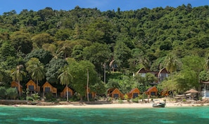Ett semesterparadis vid stranden med träbungalower, kristallklart turkost vatten och frodiga, gröna kullar i bakgrunden.