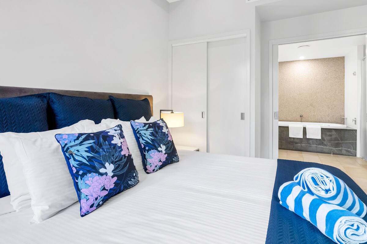 Hotellihuone, jossa on sänky, yöpöytä, lamppu, kylpyammeella varustettu kylpyhuone ja pyyhkeet.