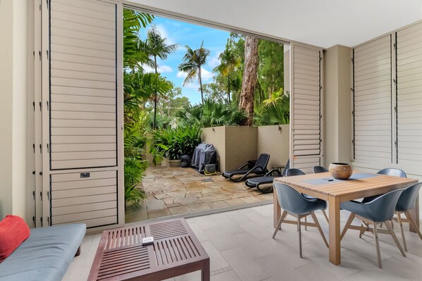 A room with a sliding glass door leading to a patio with a stone floor, outdoor seating, and lush greenery. - Elysium Triton by the Sea (Palm Cove)