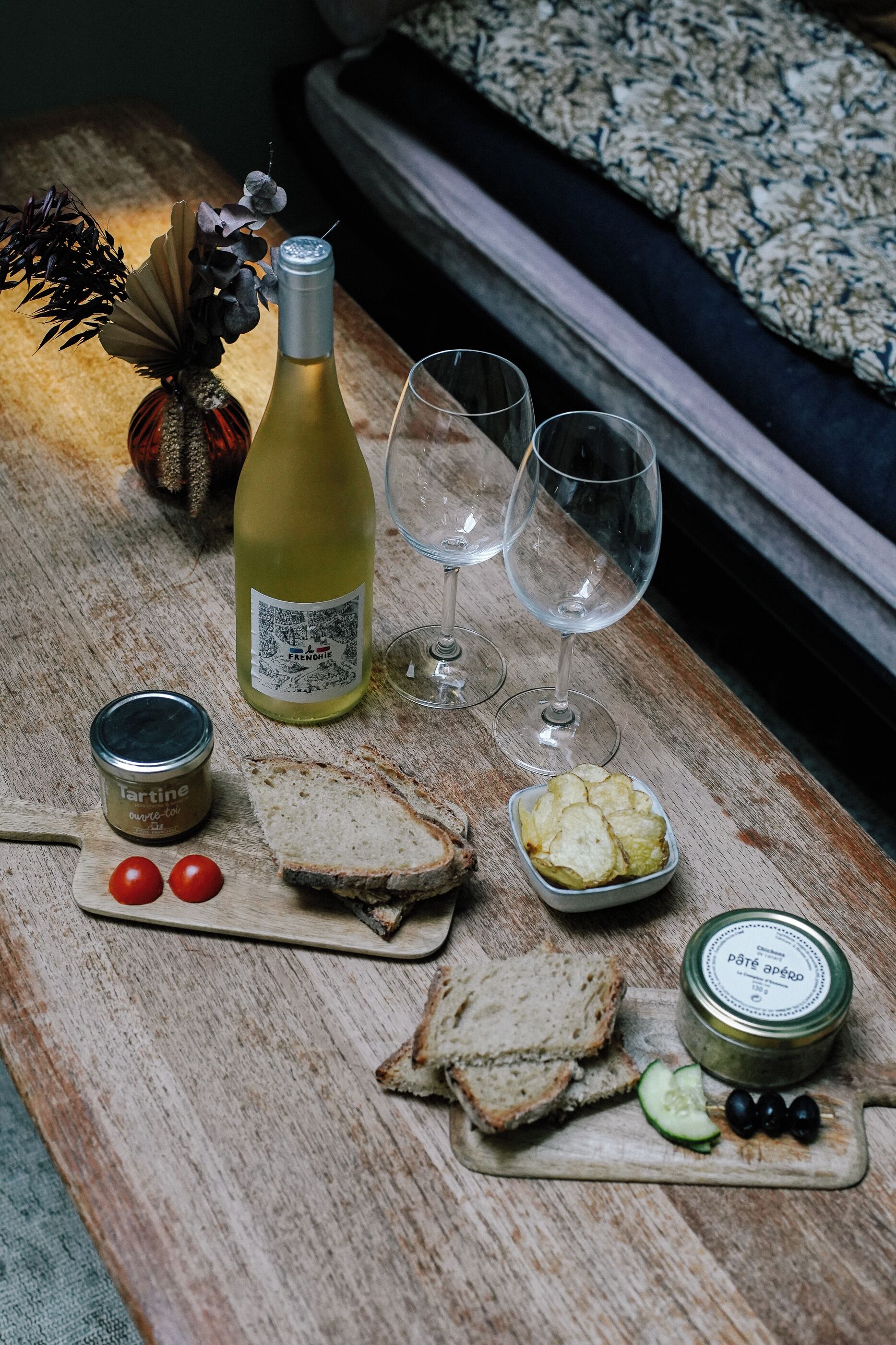 A wooden table with a bottle of wine, two glasses, a jar of tartine, a bowl of cheese, and a small container of olives.