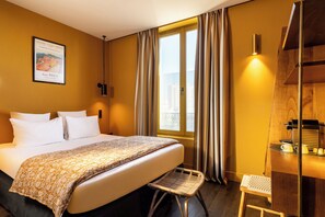 A hotel room with a bed, a small stool, a window with curtains, a wall-mounted light, and a wooden shelf with a coffee maker.