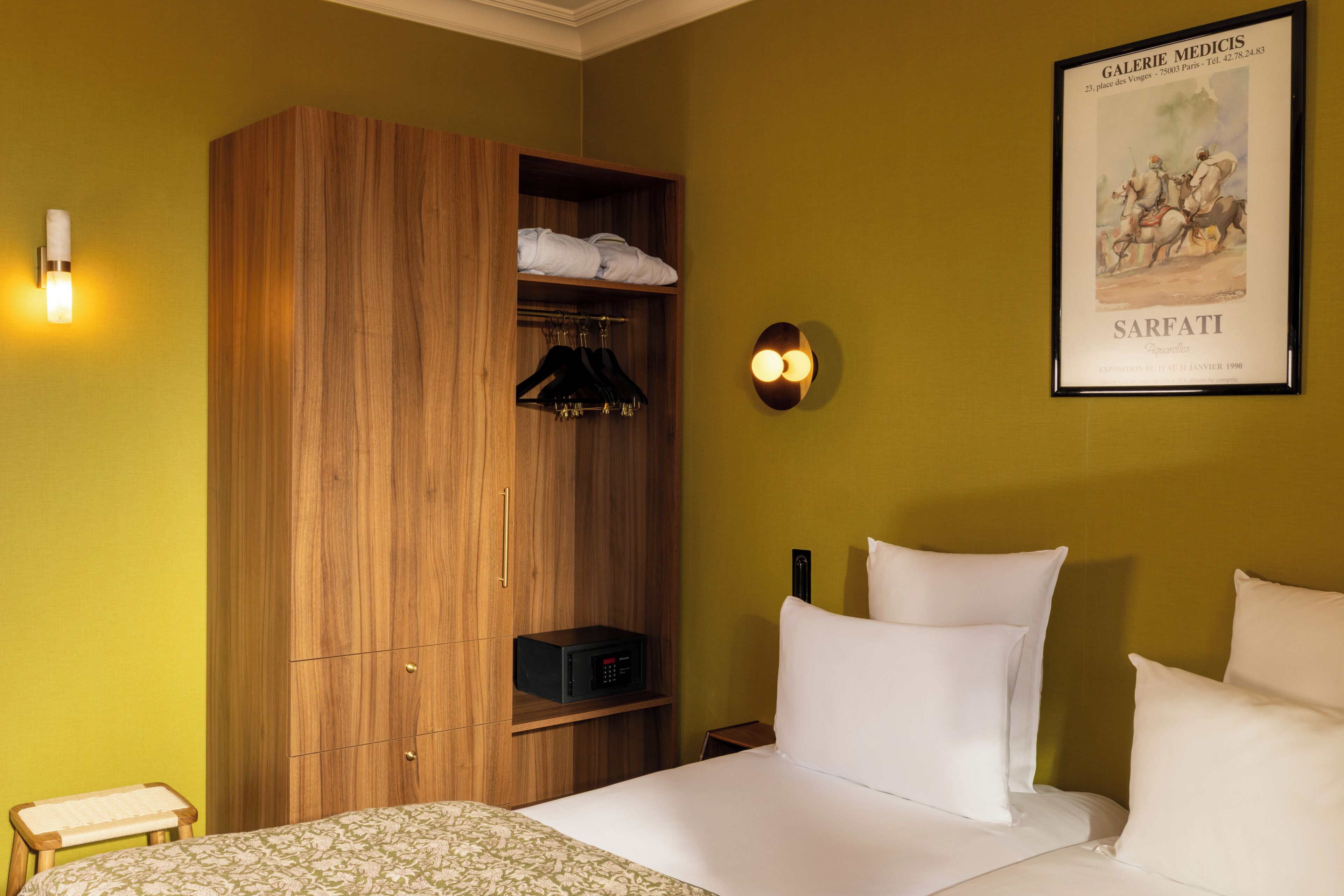 A hotel room with a bed, a wooden wardrobe, a small table, and a wall-mounted light fixture.