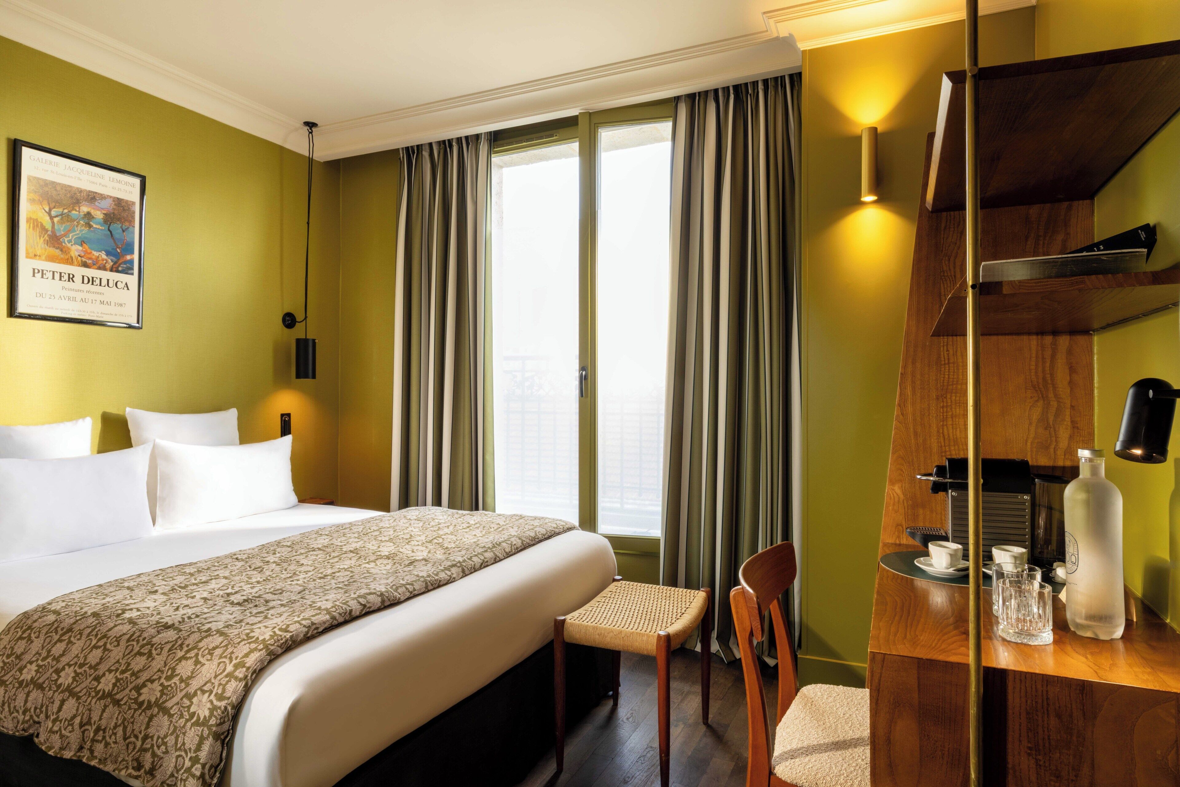 A hotel room with a bed, a chair, a desk with a coffee machine, a bottle, and a framed picture on the wall.