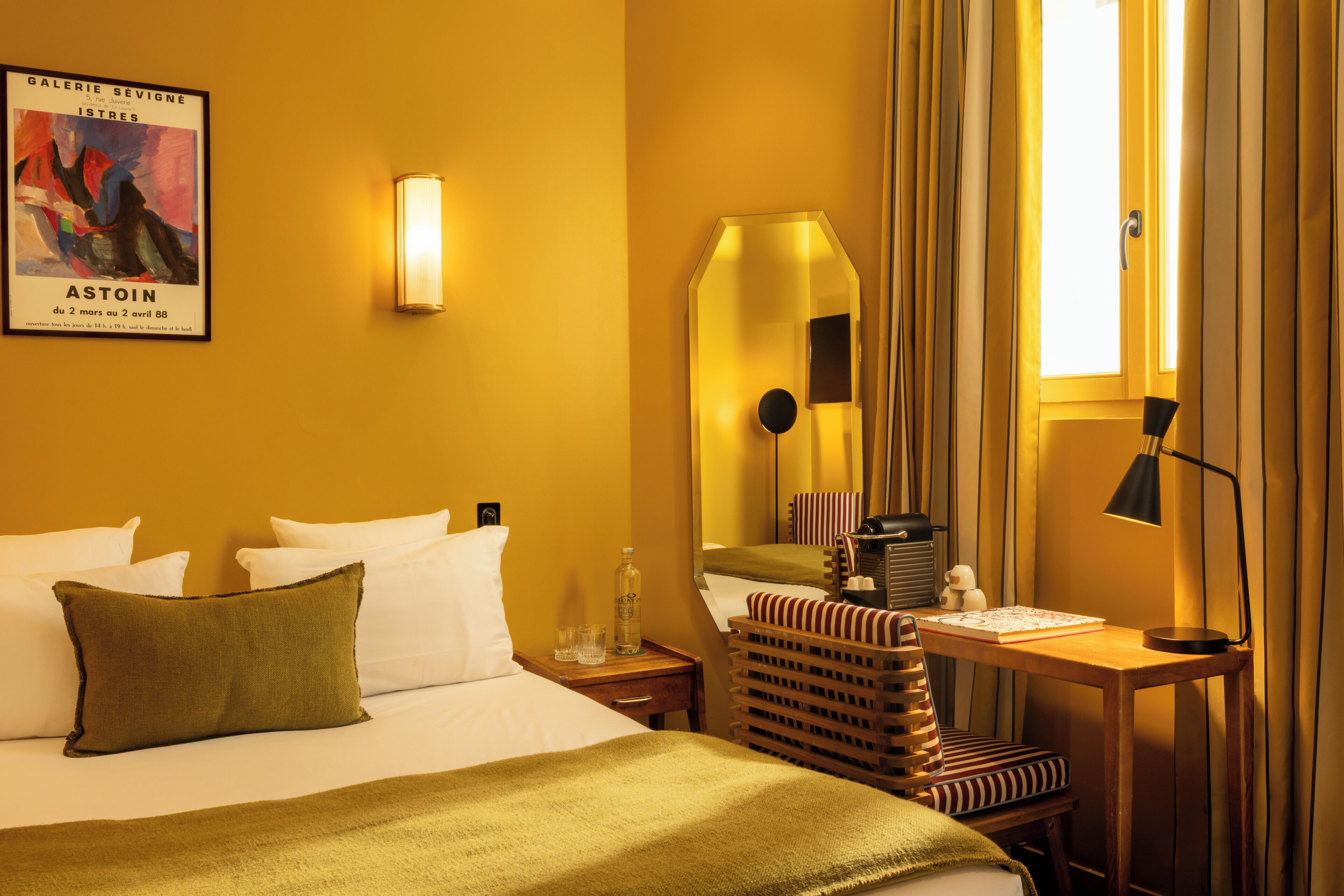 Una habitación de hotel con cama, escritorio, silla, mesita de noche, lámpara, espejo y un cartel en la pared.