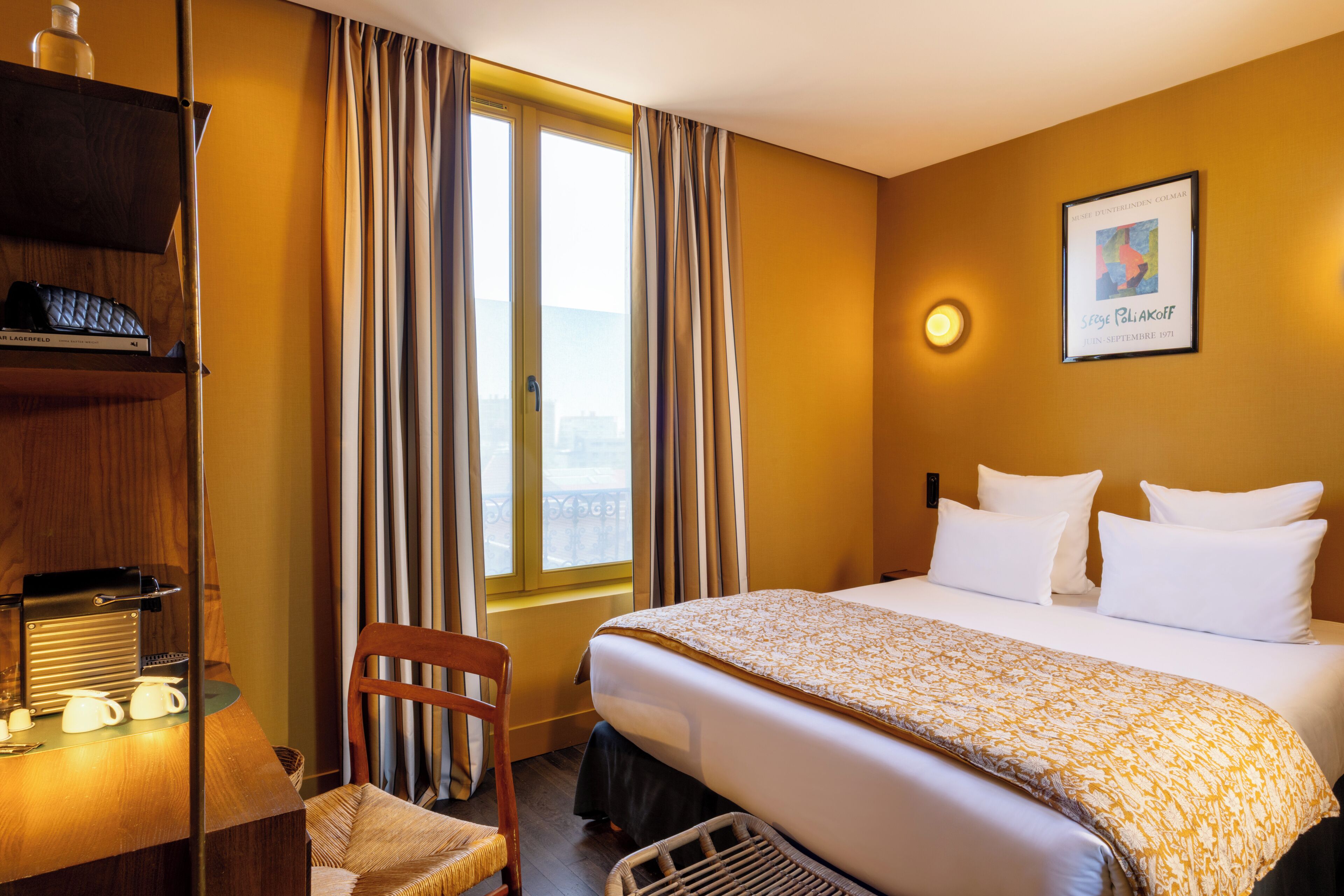 Une chambre d’hôtel avec un lit, une chaise, un bureau avec une machine à café et une fenêtre avec des rideaux.