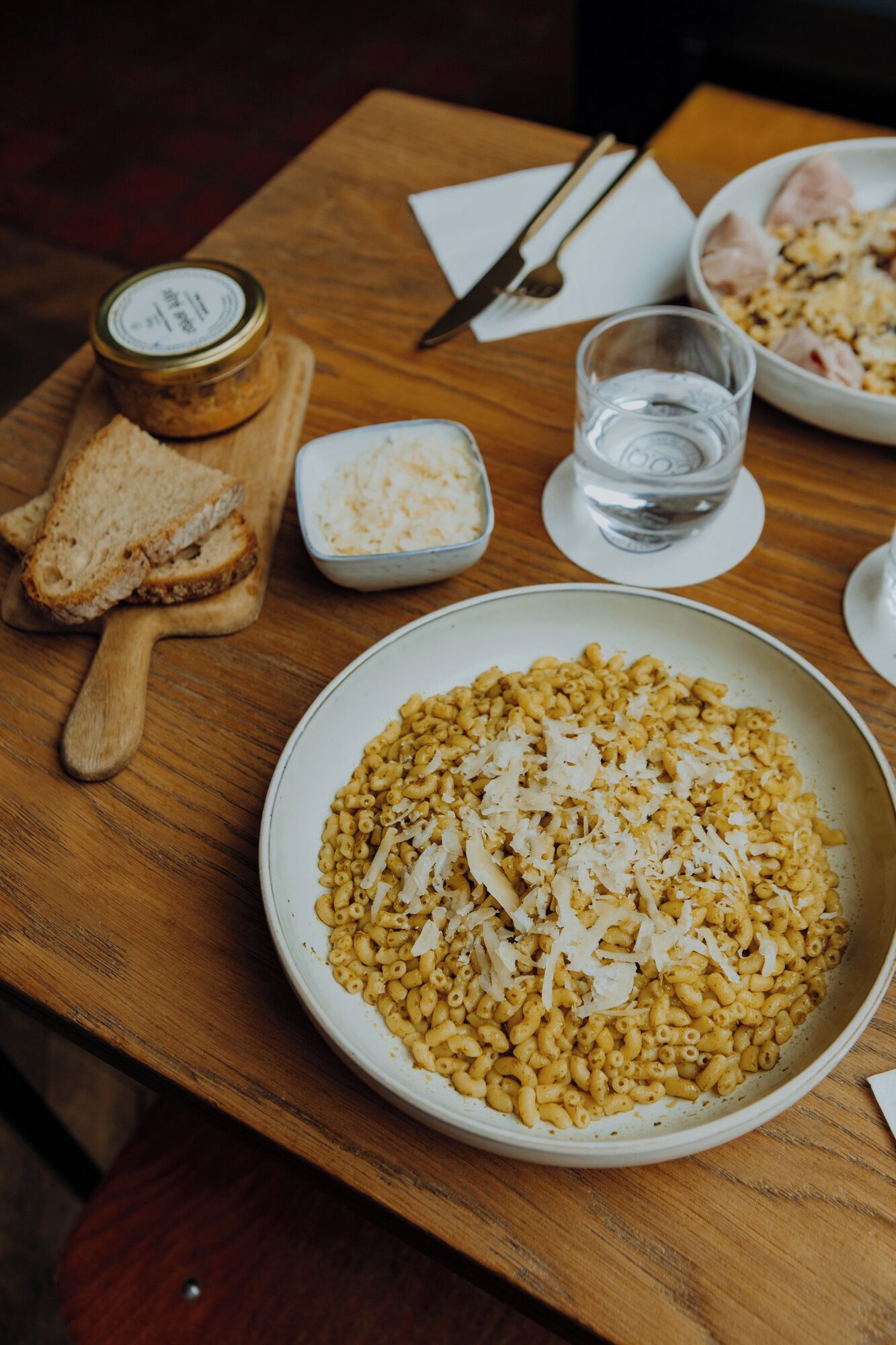 Una mesa de madera con un plato de pasta, un tarro de condimento, un recipiente pequeño de arroz, un vaso de agua y una cuchara de madera.